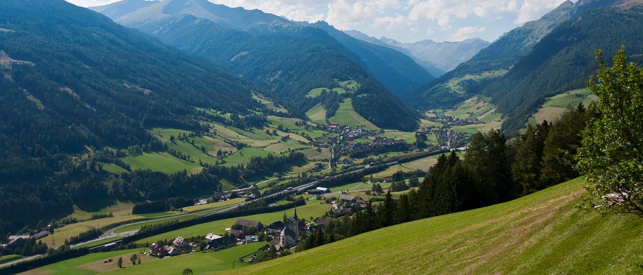 Luftaufnahme einer Bergregion mit üppigen grünen Tälern, einer kurvenreichen Straße und einem kleinen Dorf, das zwischen den Bäumen eingebettet ist.