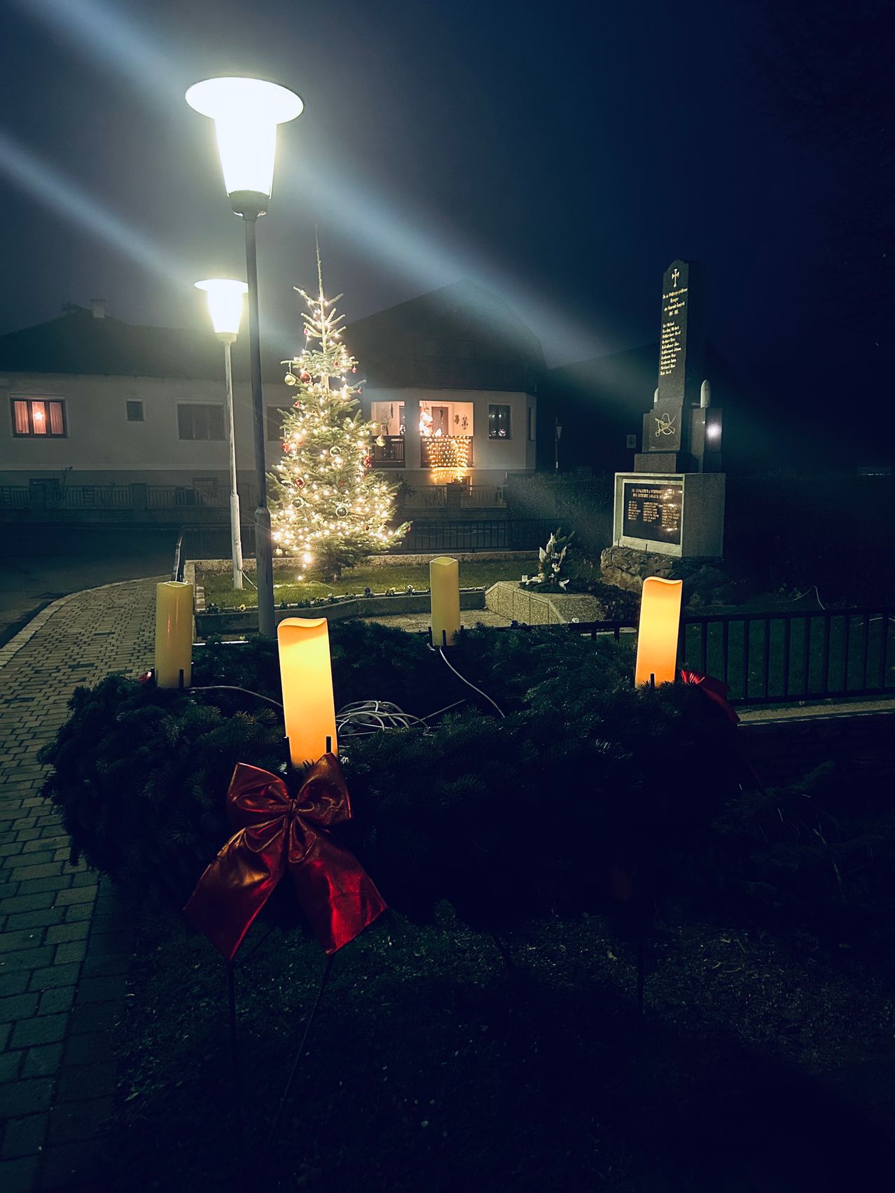 Ein Weihnachtsbaum ist vor einem Haus mit einem Gedenkstein und einem Zaun beleuchtet.