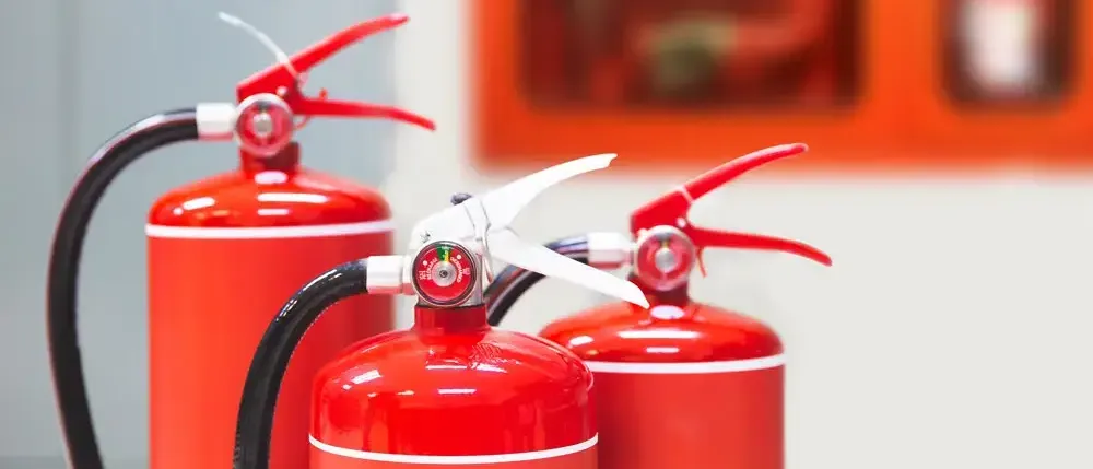 Drei rote Feuerlöscher stehen in einer Reihe an einer weißen Wand, mit einem verschwommenen orangefarbenen Gemälde im Hintergrund.