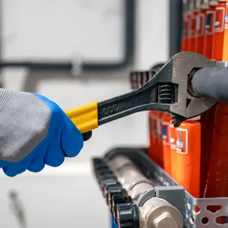 Eine Person mit Handschuhen hält einen Schraubenschlüssel und scheint etwas in einem Raum mit orangefarbenen Behältern zu reparieren.
