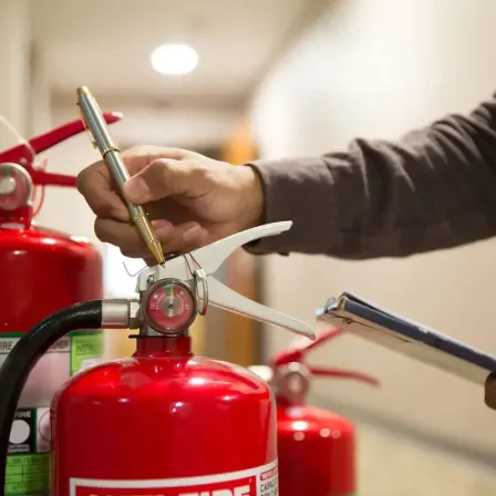 Eine Person überprüft die Druckanzeige eines roten Feuerlöschers. Sie hält einen Stift und hat einen Klemmbrett in der Nähe. Ein weiterer Feuerlöscher ist im Hintergrund sichtbar.