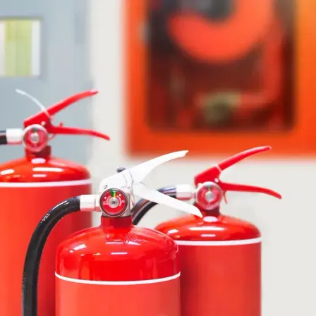 Drei rote Feuerlöscher stehen auf einer weißen Oberfläche. Die Feuerlöscher haben rote Düsen und schwarze Griffe. Dahinter ist ein unscharfes Bild eines Brandschutzposters.
