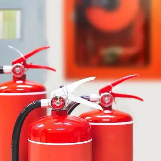 Drei rote Feuerlöscher stehen auf einer weißen Oberfläche. Die Feuerlöscher haben rote Düsen und schwarze Griffe. Dahinter ist ein unscharfes Bild eines Brandschutzposters.