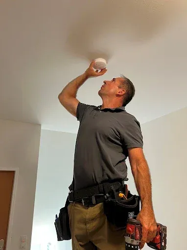 Ein Mann in einem grauen Hemd und Hose, mit einer Bohrmaschine und einem Werkzeuggürtel, installiert einen weißen Rauchmelder an der Decke.