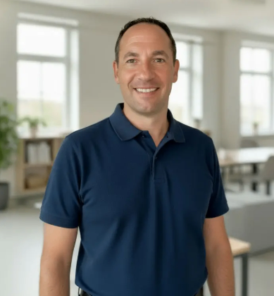 Ein Mann in einem blauen Poloshirt steht in einem hellen Raum mit weißen Wänden und großen Fenstern. Er lächelt für ein Foto.