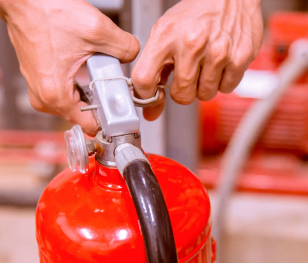Eine Person hält einen roten Feuerlöscher und ist dabei, die Sicherheitsnadel zu lösen. Der Feuerlöscher steht aufrecht auf einer weißen Oberfläche.