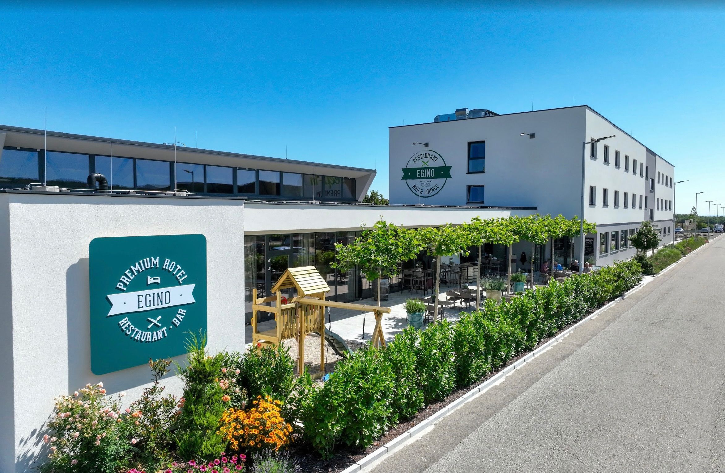 Das Premium Hotel EginO, ein modernes Gebäude, steht prominent. Ein Schild mit dem Namen und Logo des Hotels ist sichtbar, und ein Kinderspielhaus steht daneben. Das Hotel verfügt über eine großzügige Terrasse mit Tischen und Stühlen und einem üppigen Garten.