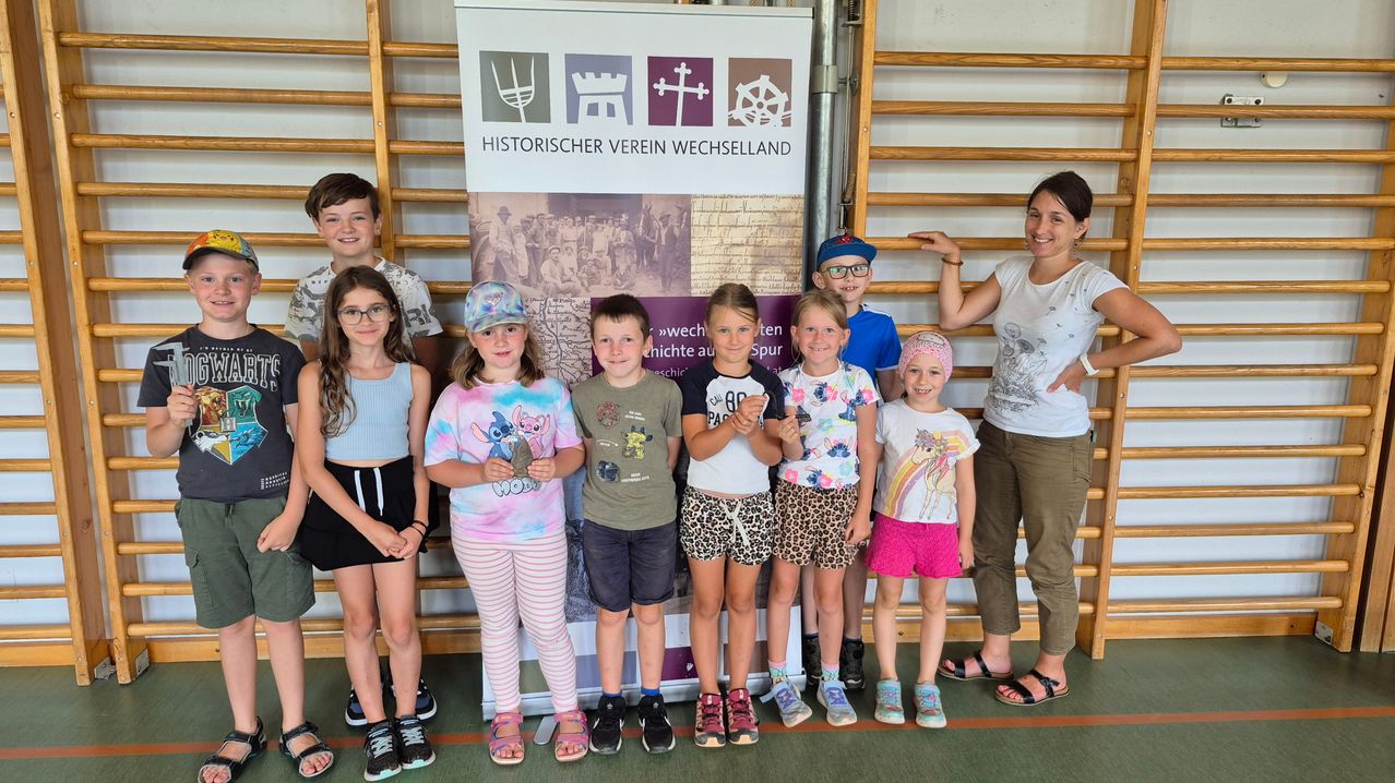 Eine Gruppe von Kindern und zwei Erwachsenen posiert für ein Foto vor einem Banner mit dem Text 'Historischer Verein Wechselland'. Sie alle lächeln und scheinen eine gute Zeit zu haben.