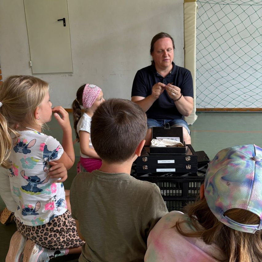 Ein erwachsener Mann unterrichtet drei Kinder über Insekten mit einer Lupe und einer Schachtel mit Exemplaren. Die Kinder sitzen auf dem Boden und hören aufmerksam zu.