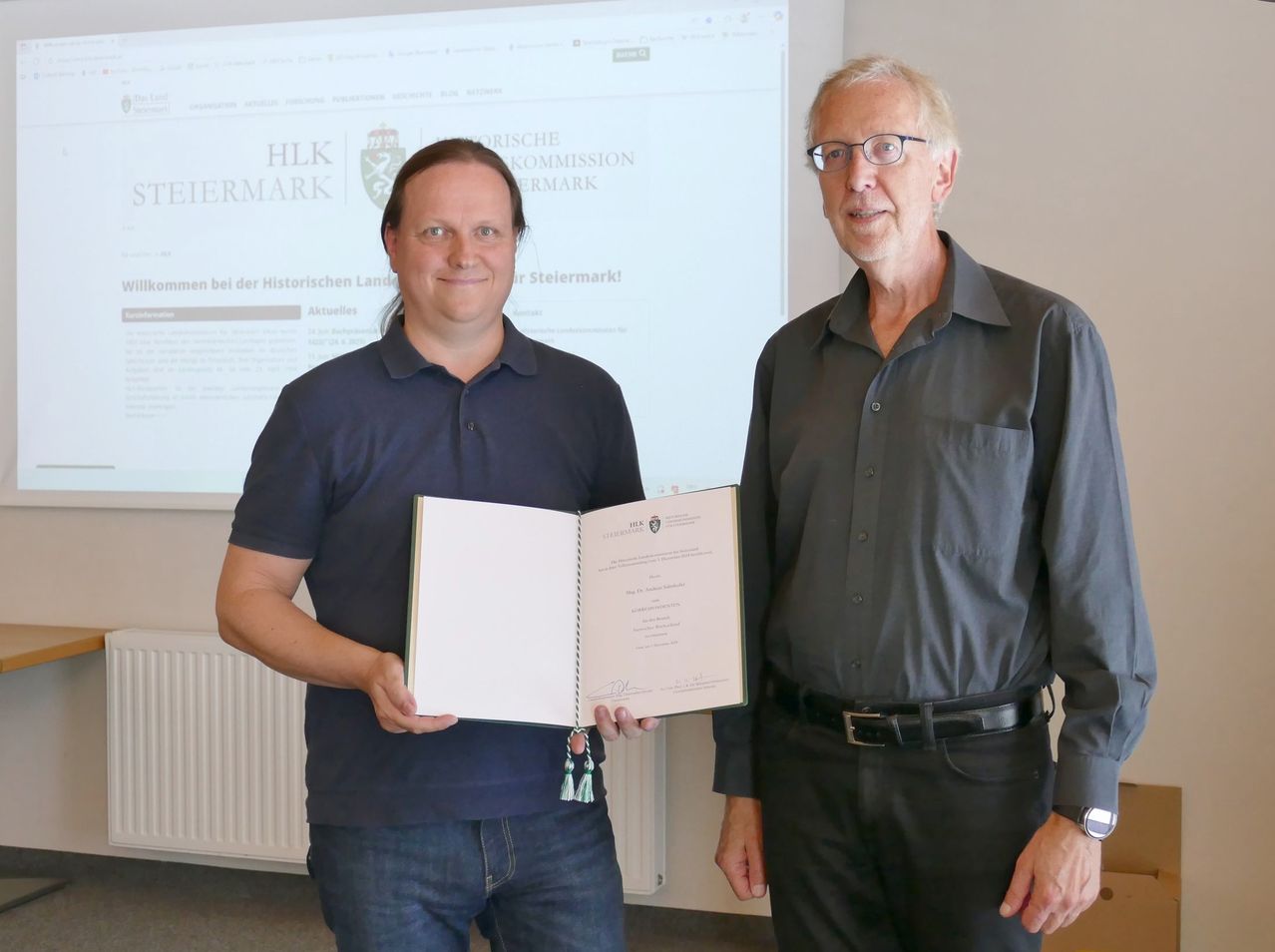 Zwei Männer stehen in einem Raum und halten ein offenes Buch. Der Mann auf der linken Seite lächelt, trägt ein dunkelblaues Hemd und Jeans. Der Mann auf der rechten Seite trägt eine Brille, ein graues Hemd und einen schwarzen Gürtel. Hinter ihnen ist ein Bildschirm, der eine Webseite anzeigt.