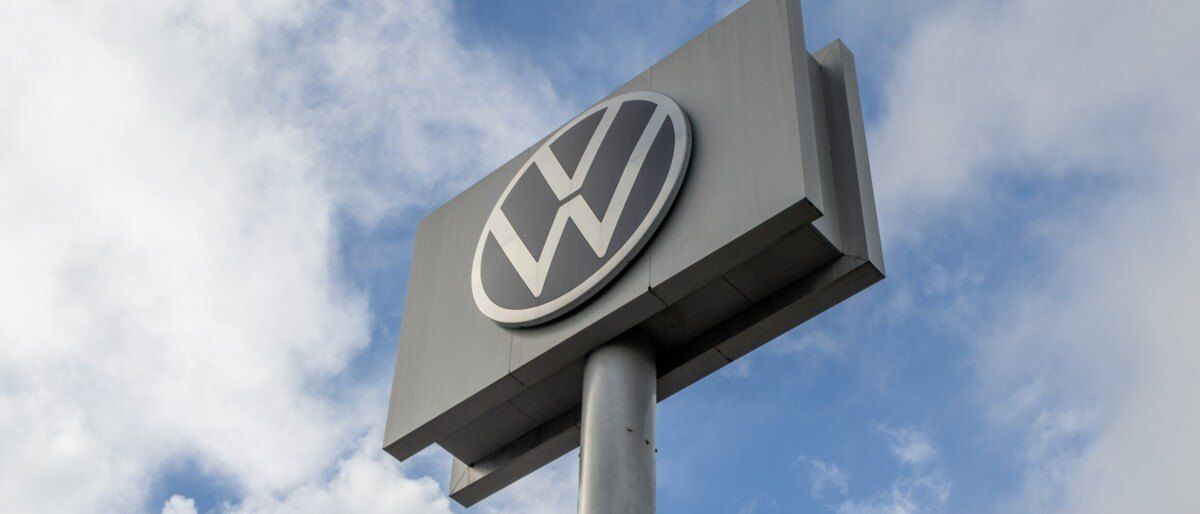 Ein VW-Logo ist auf einem grauen Schild angebracht, vor einem blauen Himmel mit weißen Wolken.