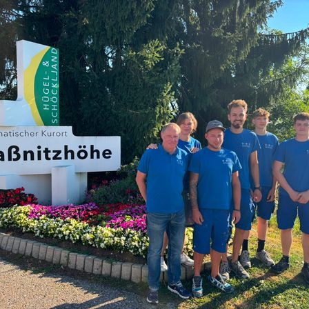 Eine Gruppe von Menschen in blauen Uniformen steht vor einem Schild, auf dem 'Nationaler Kurort Bnitzhoehe' steht. Sie lächeln und scheinen für ein Foto zu posieren. Hinter ihnen befinden sich Blumen und Bäume.