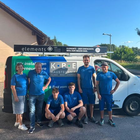 Eine Gruppe von Menschen in blauen Uniformen posiert vor einem Service-Van mit den Texten 'elements' und 'KBE Haustechnik' oben. Sie stehen auf einem Kiesweg mit einem Haus und Bäumen im Hintergrund.