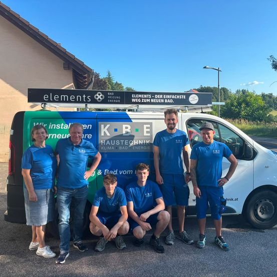 Eine Gruppe von Menschen in blauen Uniformen posiert vor einem Service-Van mit den Texten 'elements' und 'KBE Haustechnik' oben. Sie stehen auf einem Kiesweg mit einem Haus und Bäumen im Hintergrund.