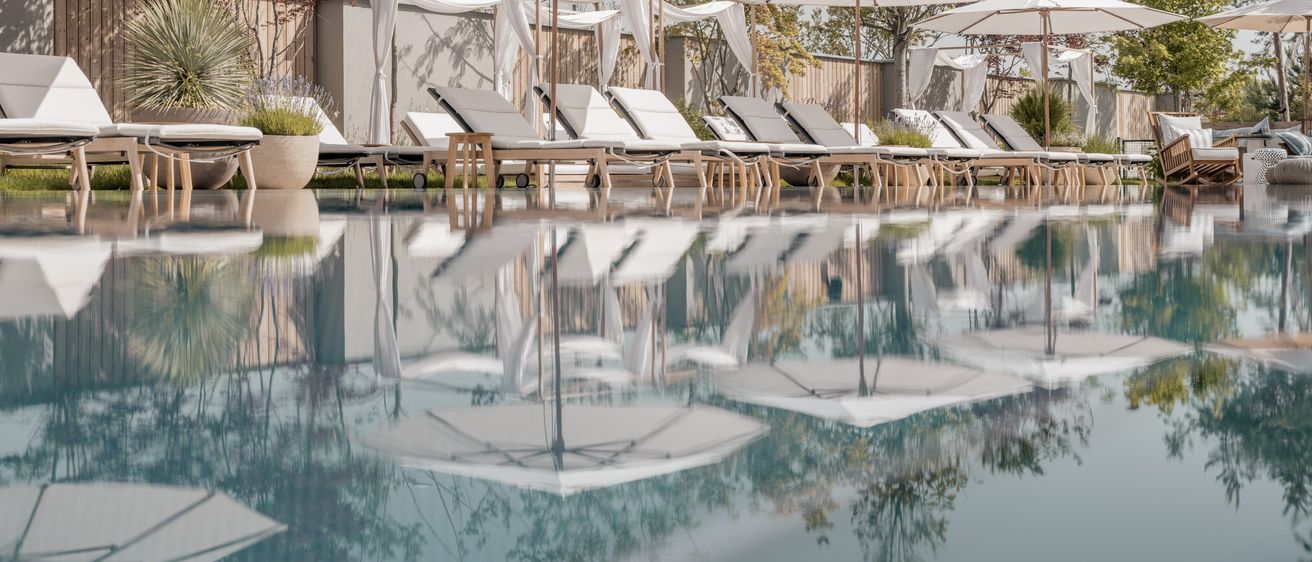 Ein Außenpoolbereich mit mehreren weißen Liegestühlen und Sonnenschirmen, die sich im Wasser spiegeln. Der Pool ist von einem Holzzaun umgeben und im Hintergrund befinden sich Bäume.