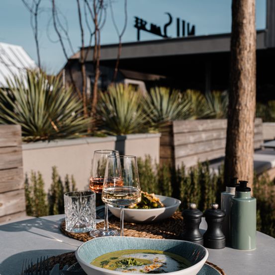 Ein Tisch für das Essen im Freien mit einer Schüssel Suppe, Weingläsern und Gewürzen auf einer grauen Tischdecke. Im Hintergrund befindet sich ein Gebäude mit einem Baum und Pflanzen.
