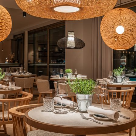 Ein Essbereich in einem Restaurant mit runden Holztischen, Stühlern und Pendelleuchten. Die Tische haben Pflanzen, Gläser und Besteck.