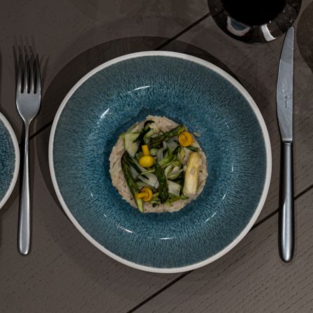 Nahaufnahme eines blauen Keramiktellers, gefüllt mit einem bunten Gemüsegericht bestehend aus Spargel, Artischocken und Zitrone. Eine Gabel und ein Messer stehen neben dem Teller, mit einem Weinglas im Hintergrund.