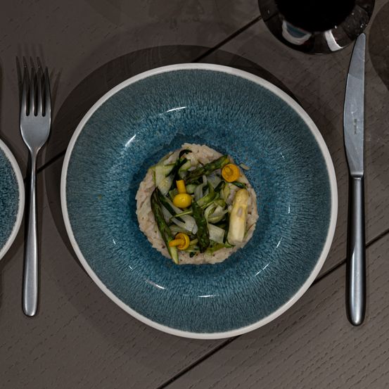 Nahaufnahme eines blauen Keramiktellers, gefüllt mit einem bunten Gemüsegericht bestehend aus Spargel, Artischocken und Zitrone. Eine Gabel und ein Messer stehen neben dem Teller, mit einem Weinglas im Hintergrund.