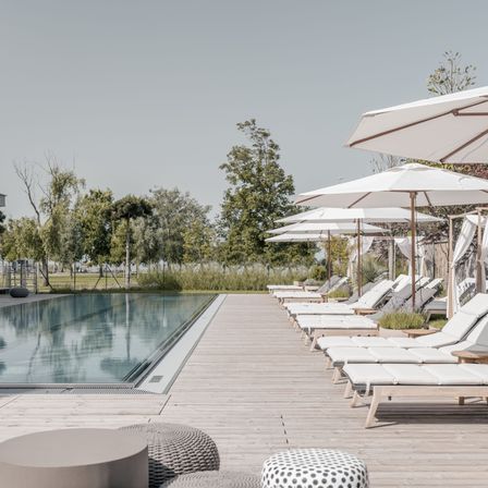 Eine Reihe von Liegestühlen mit weißen Kissen und Sonnenschirmen auf einer Holzterrasse neben einem Schwimmbad mit Spiegelungen von Bäumen und Himmel.