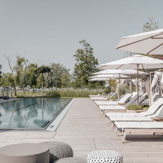 Eine Reihe von Liegestühlen mit weißen Kissen und Sonnenschirmen auf einer Holzterrasse neben einem Schwimmbad mit Spiegelungen von Bäumen und Himmel.