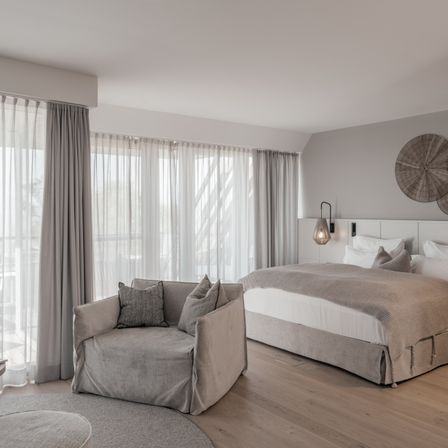 Ein elegantes Schlafzimmer mit einem großen Bett und einem gemütlichen Sessel. Der Raum hat weiße Wände, Holzböden und graue Vorhänge.