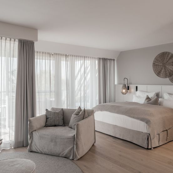 Ein elegantes Schlafzimmer mit einem großen Bett und einem gemütlichen Sessel. Der Raum hat weiße Wände, Holzböden und graue Vorhänge.