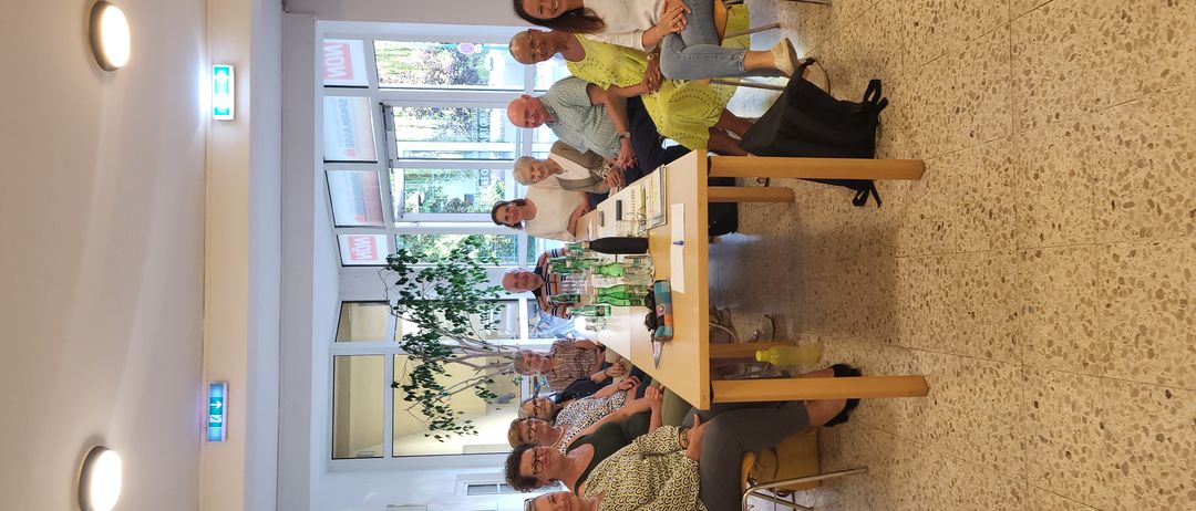 Eine Gruppe von Menschen sitzt in einem Raum mit einem Tisch, Stühlen und einer Pflanze. Der Raum hat große Fenster und ein Schild mit der Aufschrift 'JETZT'.