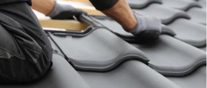 Eine Person in schwarzer Kleidung und Handschuhen montiert graue Dachziegel auf einer hölzernen Dachstruktur.