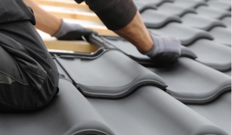 Eine Person in schwarzer Kleidung und Handschuhen montiert graue Dachziegel auf einer hölzernen Dachstruktur.