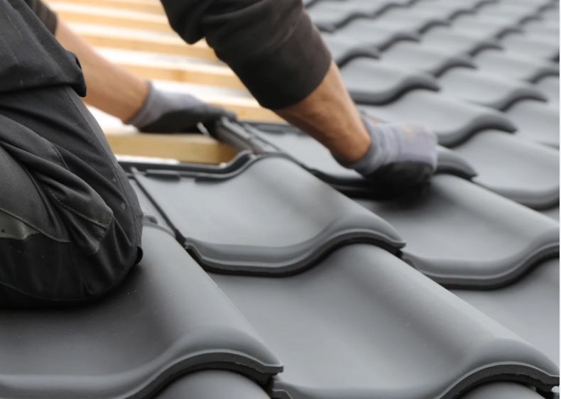 Eine Person in schwarzer Kleidung und Handschuhen montiert graue Dachziegel auf einer hölzernen Dachstruktur.