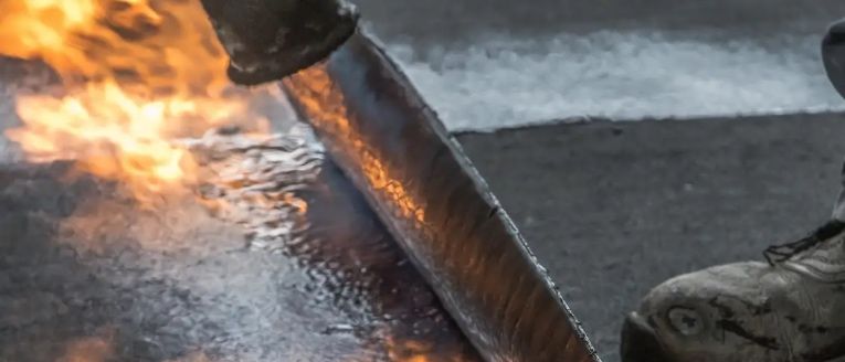 Eine Nahaufnahme eines brennenden Planens mit einem Metallrinnen in der Nähe. Die Plane ist vollständig von Flammen umhüllt, mit aufsteigendem Rauch und einer nassen Oberfläche darunter. Ein Schuh eines Menschen ist in der Ecke sichtbar, was darauf hindeutet, dass sie in der Nähe sind.