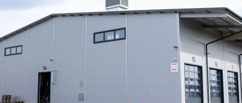 Ein großes, graues Industriegebäude mit Dachlüftern und mehreren Türen, nummeriert 14, 15, 16 und 17, befindet sich in einem offenen Bereich unter einem bewölkten Himmel.