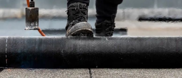 Eine Person in Schutzausrüstung benutzt einen Brenner, um ein schwarzes Dachmaterial auf einem Dach zu versiegeln.