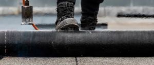 Eine Person in Schutzausrüstung benutzt einen Brenner, um ein schwarzes Dachmaterial auf einem Dach zu versiegeln.