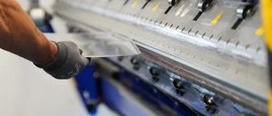 Eine Person mit Handschuhen benutzt eine Metallbiegemaschine mit einem glänzenden Metallblech, vor einem weißen Hintergrund.