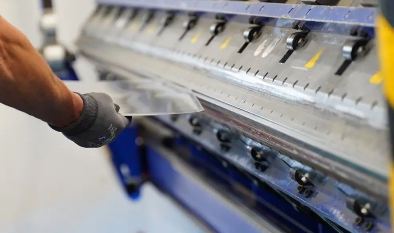 Eine Person mit Handschuhen benutzt eine Metallbiegemaschine mit einem glänzenden Metallblech, vor einem weißen Hintergrund.