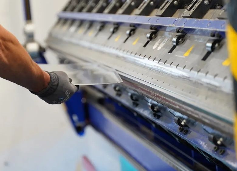 Eine Person mit Handschuhen benutzt eine Metallbiegemaschine mit einem glänzenden Metallblech, vor einem weißen Hintergrund.