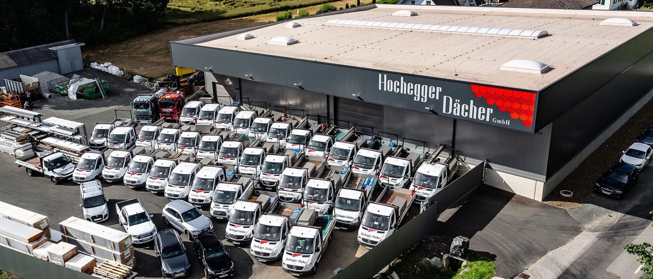 Eine Luftaufnahme eines großen Parkplatzes mit mehreren geparkten Autos und Lieferwagen. Ein Gebäude mit den Worten Hochegger Dacher GmbH ist sichtbar.