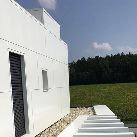 Ein modernes weißes Gebäude mit einem kleinen Fenster und Stufen, die zu einem Rasenfeld mit Bäumen im Hintergrund führen.