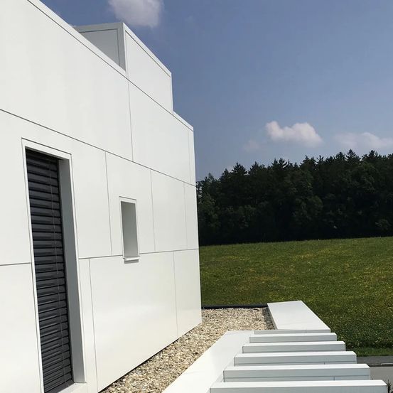 Ein modernes weißes Gebäude mit einem kleinen Fenster und Stufen, die zu einem Rasenfeld mit Bäumen im Hintergrund führen.