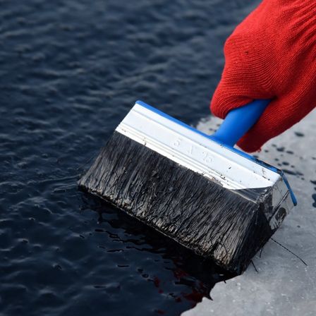 Eine Person in roten Handschuhen verwendet einen Pinsel, um eine dunkle Substanz auf eine feuchte Oberfläche aufzutragen. Der Pinsel hat einen blauen Griff und eine weiße Umrandung.