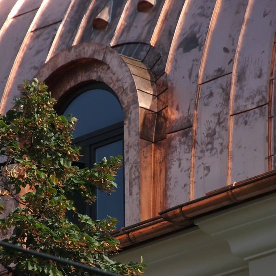 Eine Nahaufnahme eines Kupferdaches und einer Dachrinne eines Gebäudes mit einem Fenster und einem Baumzweig im Vordergrund.