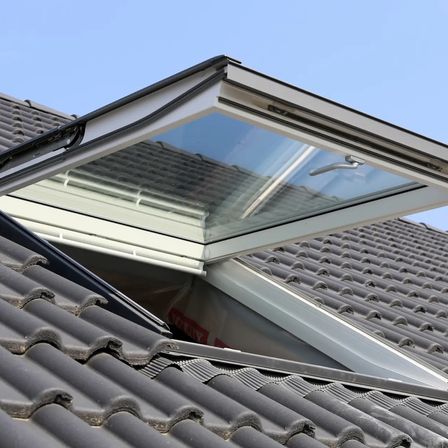 Ein Dach mit einem offenen weißen Fenster an einem klaren blauen Himmel. Das Fenster ist nach oben geneigt und hat einen silbernen Griff.