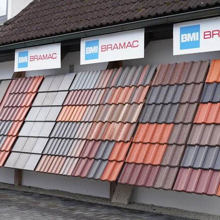 Eine Reihe verschiedener farbiger Dachziegel, die vor einem Gebäude mit BMI Bramac-Schildern ausgestellt sind.