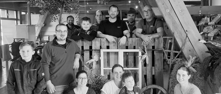 Eine Gruppe von Menschen, einschließlich Erwachsener und Kindern, posiert für ein Foto in einer Scheune. Sie sind von Holzkonstruktionen und Pflanzen umgeben.