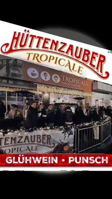 Eine Menschenmenge versammelt sich vor einem festlich dekorierten Café mit einem Schild, das Huttenzauber Tropicale anzeigt, mit einem tropisch gestalteten Café und Gelato. Das Banner wirbt für Glühwein und Punsch.