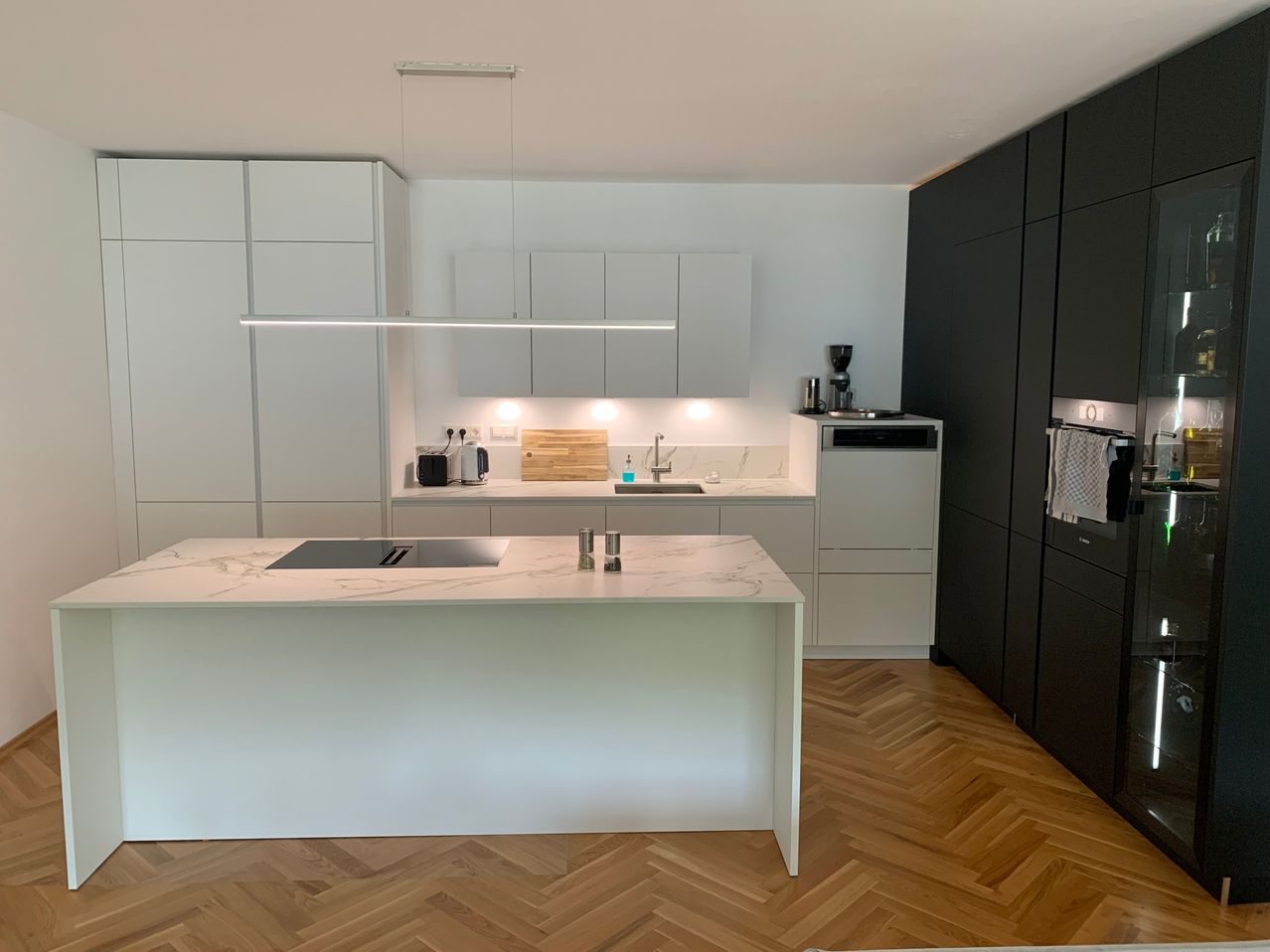 Moderne Küche mit weißen Schränken, Marmorarbeitsplatte, schwarzem Induktionsherd und Holzboden. Geräte umfassen eine Kaffeemaschine und einen Geschirrspüler.