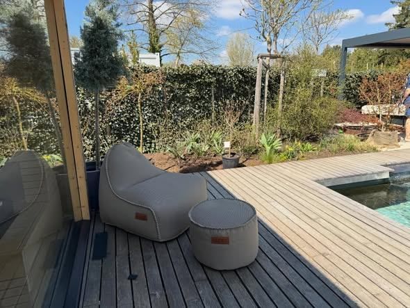 A gray bean bag chair and ottoman on a wooden deck by a pool, with a hedge and trees in the background.