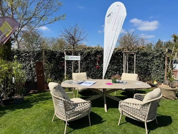 A table and chairs are set up in a garden with a white flag. The garden is surrounded by a hedge with a sign on it. There are potted plants and trees in the area.